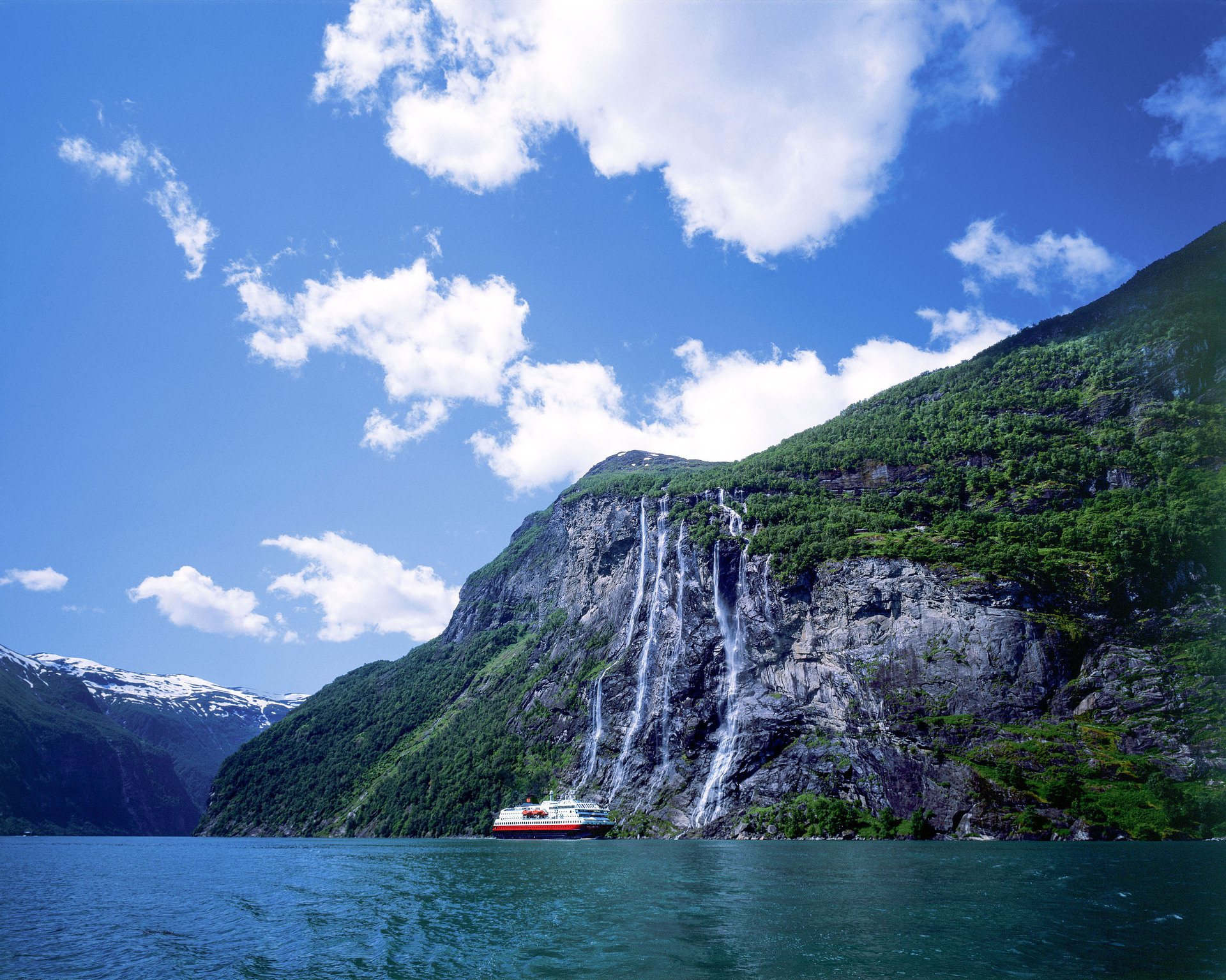 Bilden visar Hurtigrutens fartyg på håll framför vattenfallet sju systrar i Geirangerfjorden