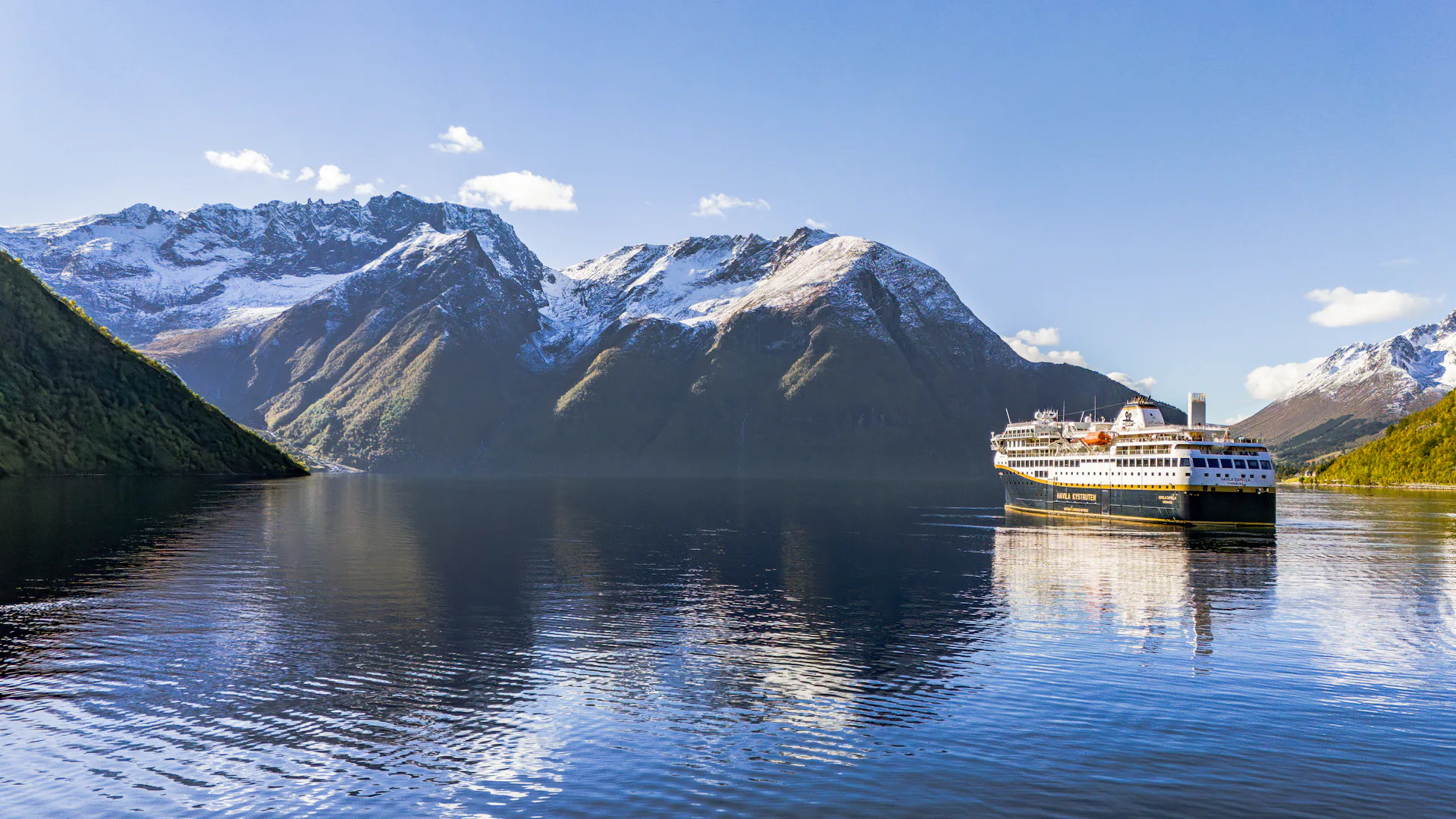 Bilden visar Havilas fartyg som seglar en vacker dag in i Hjörundfjorden. Det är strålande sol och lite snö kvar på bergstopparna.