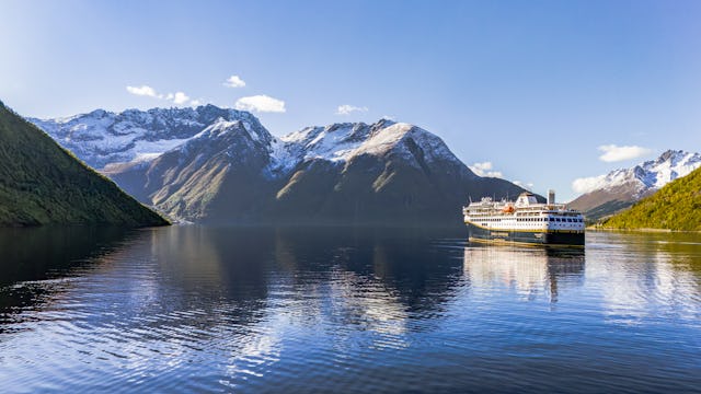 Bilden visar Havilas fartyg som seglar en vacker dag in i Hjörundfjorden. Det är strålande sol och lite snö kvar på bergstopparna.