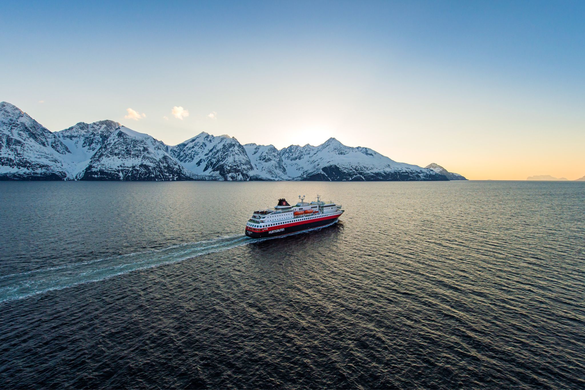 Bilden visar Hurtigrutens fartyg till havs med snötäckta berg i bakgrunden. Fartyget seglar mot solnedgången.