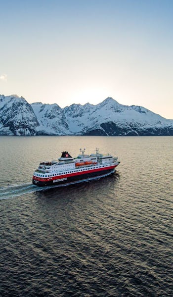 Bilden visar Hurtigrutens fartyg till havs med snötäckta berg i bakgrunden. Fartyget seglar mot solnedgången.