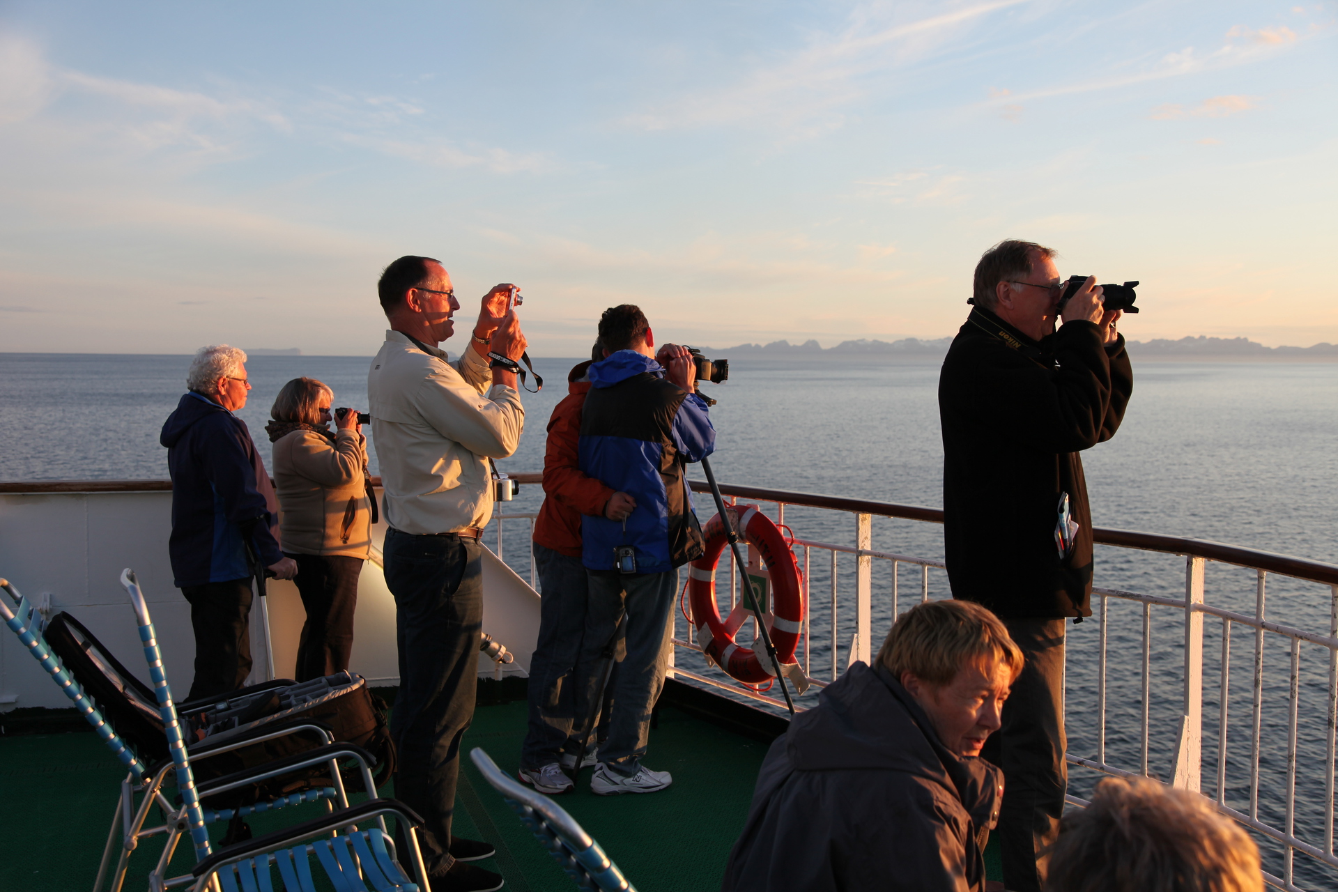 Bilden visar en grupp människor på däck där de tittar på och fotar solnedgången.