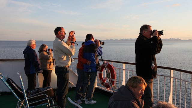 Bilden visar en grupp människor på däck där de tittar på och fotar solnedgången.