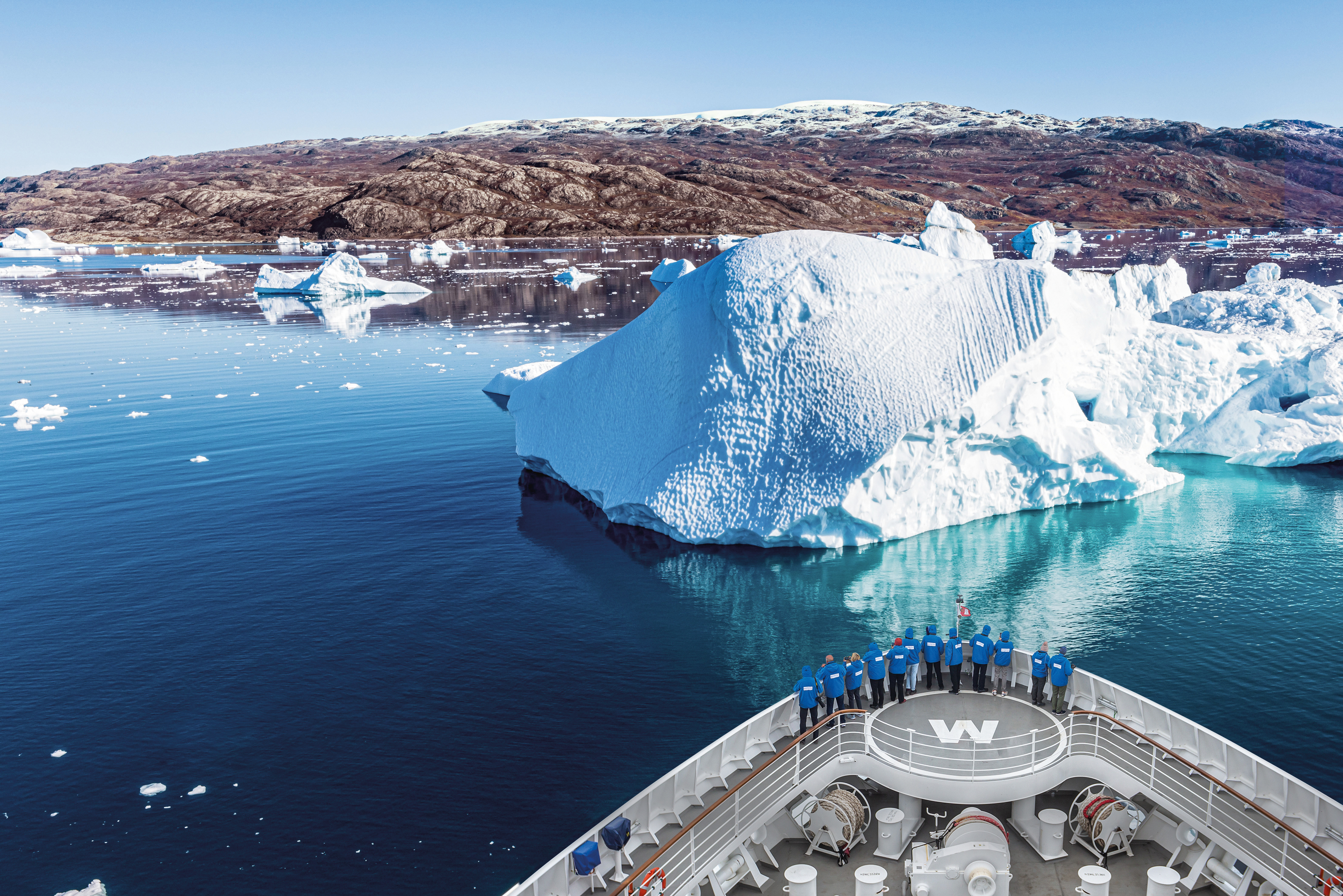 Bilden visar fören på ett av Hapag-Lloyds fartyg där personer står och tittar på isberget lite längre fram. Bilden är taget på Grönland.