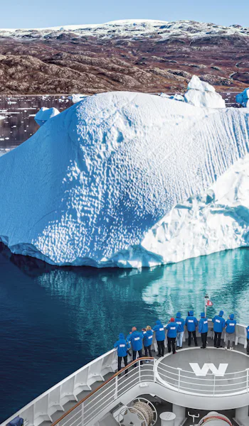 Bilden visar fören på ett av Hapag-Lloyds fartyg där personer står och tittar på isberget lite längre fram. Bilden är taget på Grönland.