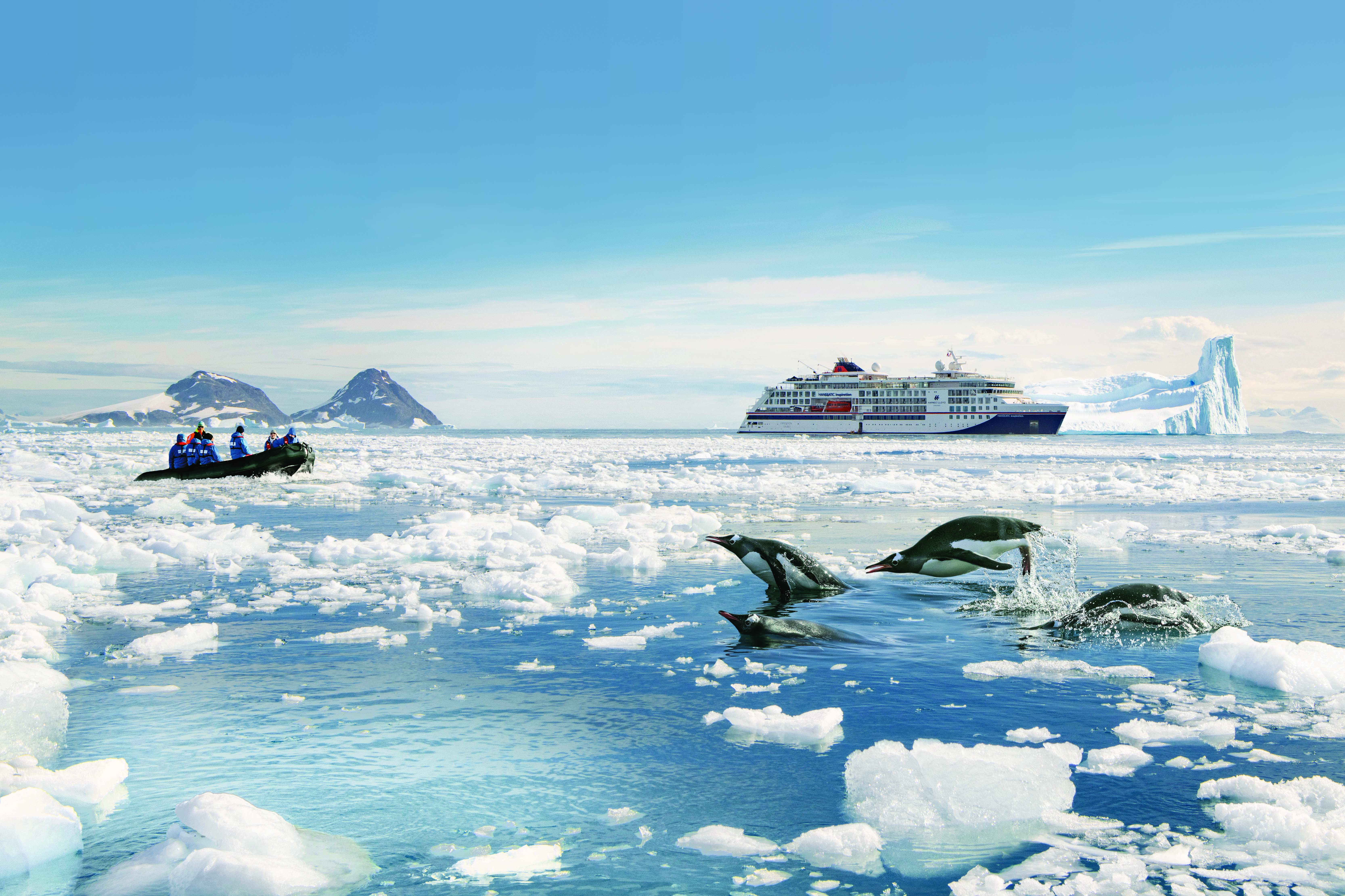 Bilden visar Hapag-Lloyds fartyg i Antarktis. Pingviner hoppar i vattnet framför fartyget.